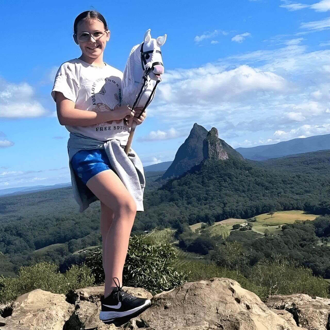 A person stands on a rocky hilltop holding a toy horse, with green hills and a distinctive steep hill in the background under a partly cloudy sky.