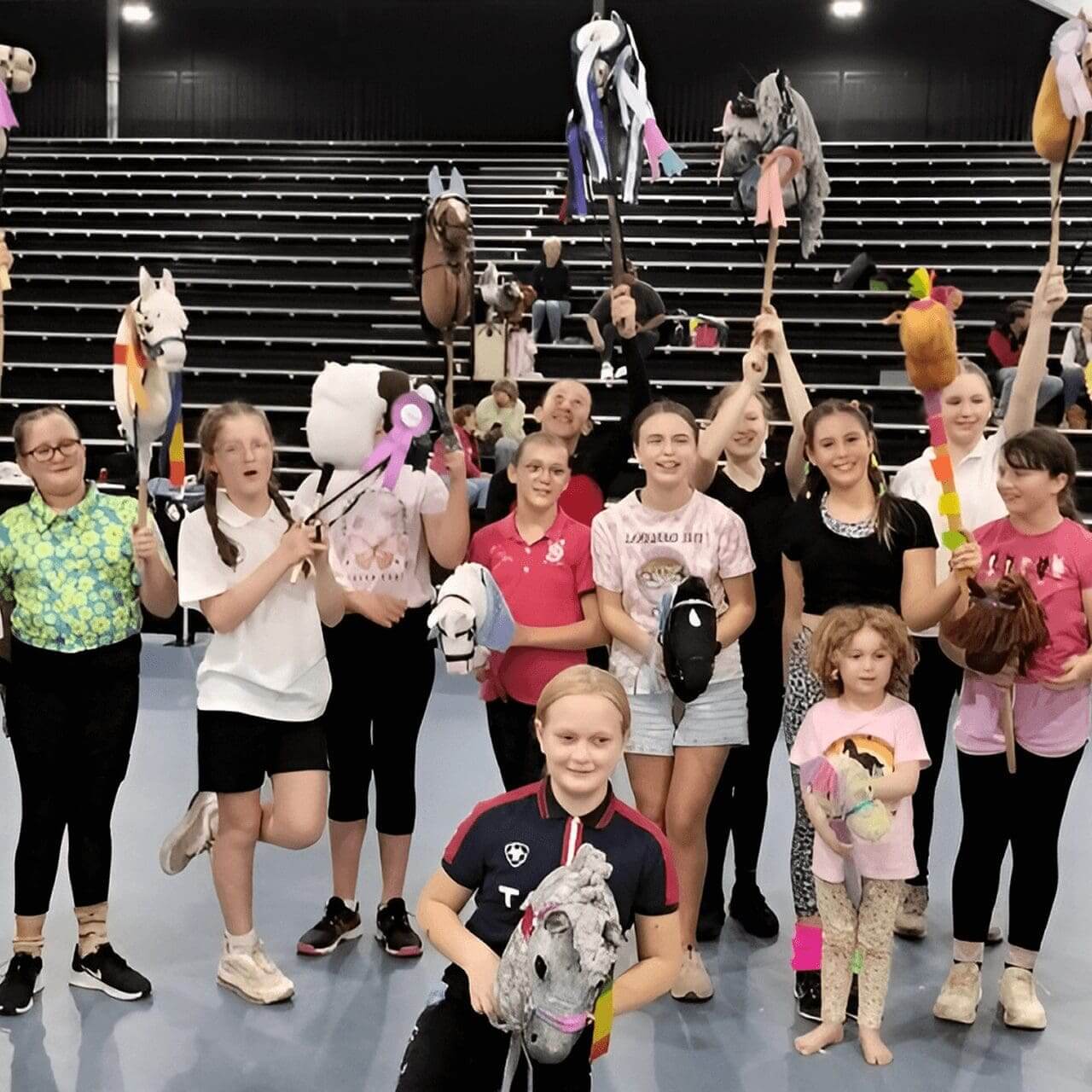 A group of children holds hobby horses, posing indoors in front of empty bleachers. They appear excited and are dressed casually.