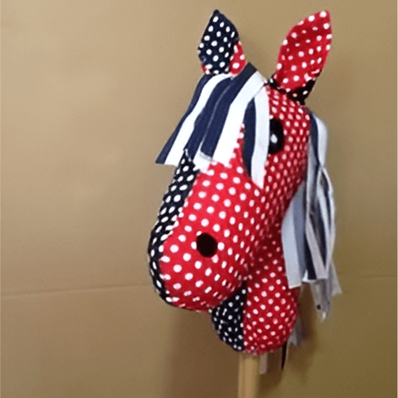 A red and white polka-dotted plush horse head with a stick, featuring blue and white striped mane and ears on a beige background.