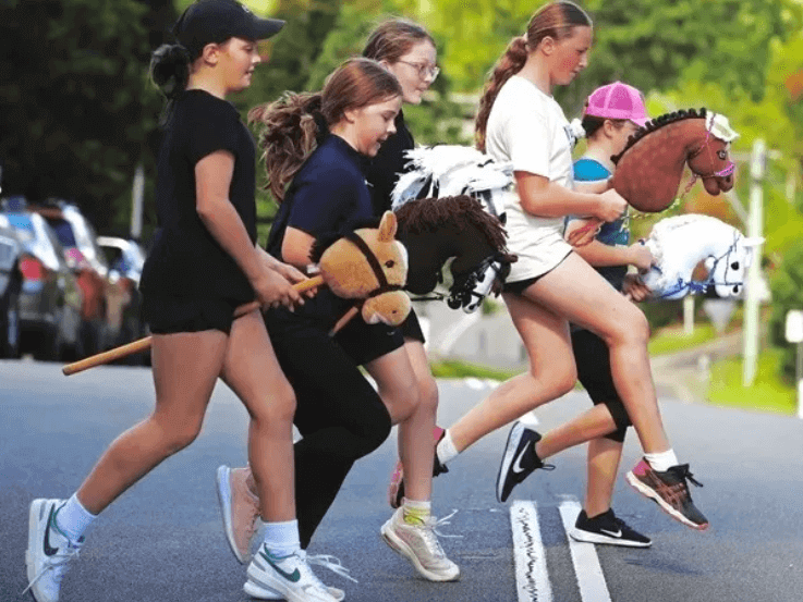 Young girls racing with hobby horses on a track.