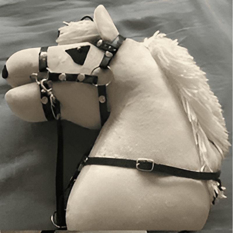 Side view of a white toy horse head with a black bridle and reins, featuring a mane of white yarn against a plain background.