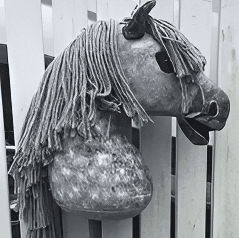 A black and white image of a hobby horse head with yarn mane, mounted on a stick, set against a wooden fence background.