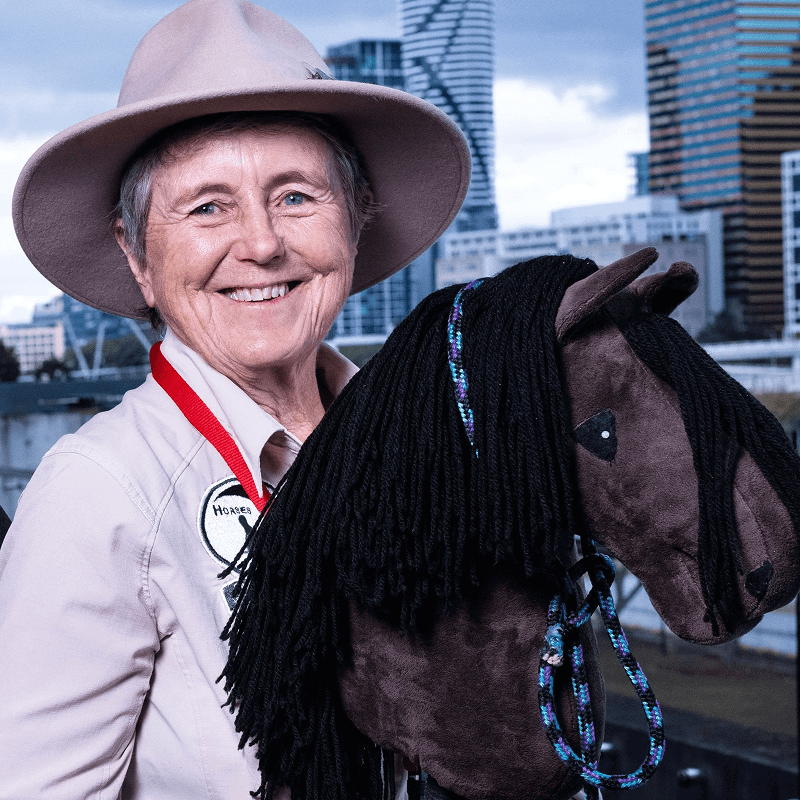 Person wearing a hat and a badge smiles while holding a toy horse with city buildings in the background.