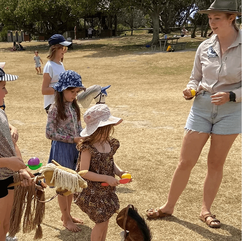 Children with hobby horses engaging in an outdoor activity with a woman holding rubber balls, on a grassy field.