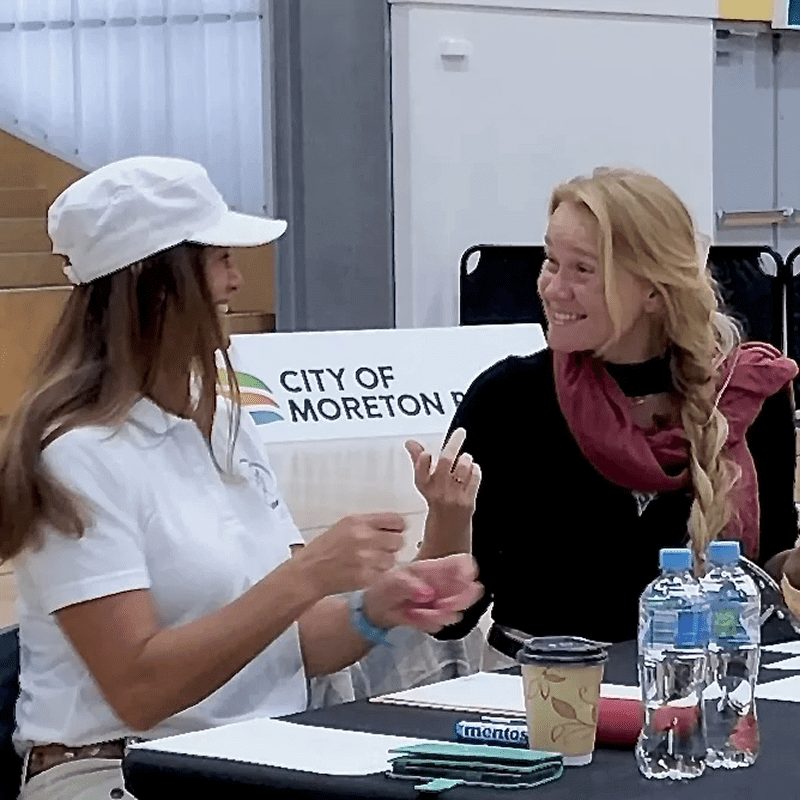 Two women sit at a table with a "City of Moreton" banner behind them. One wears a white cap and the other a scarf. A Mentos pack, water bottles, and coffee cup are on the table.