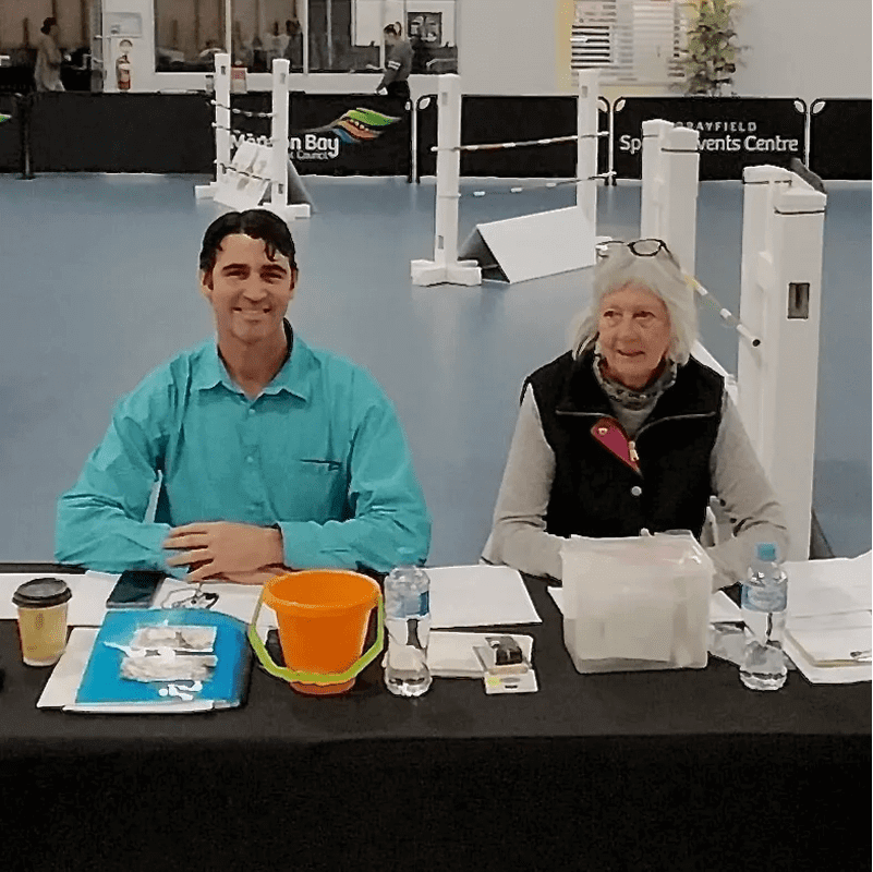 Two people sitting behind a table with papers, a coffee cup, and a bucket on it. They're in an indoor sports venue with white structures in the background.
