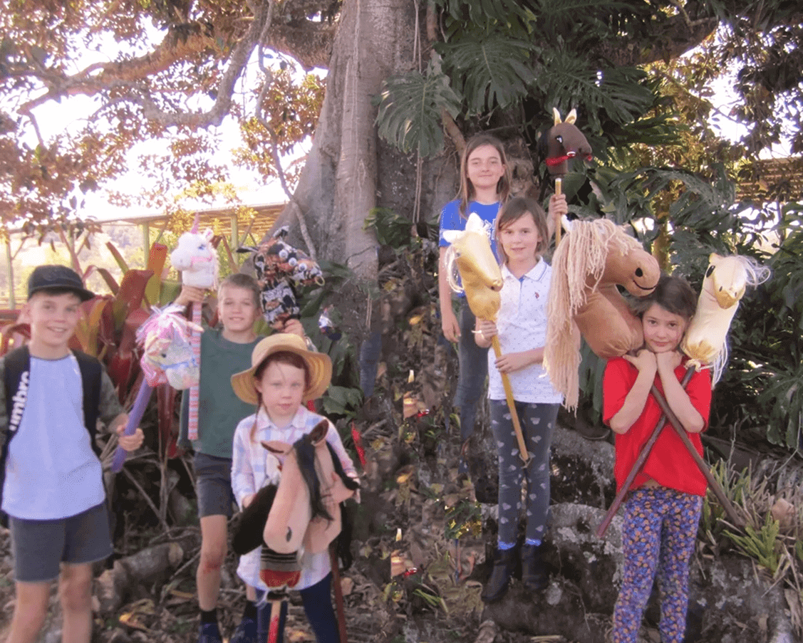 A group of children hold hobby horses and stand on a tree in a garden, posing for a photo under a tree with lush greenery.