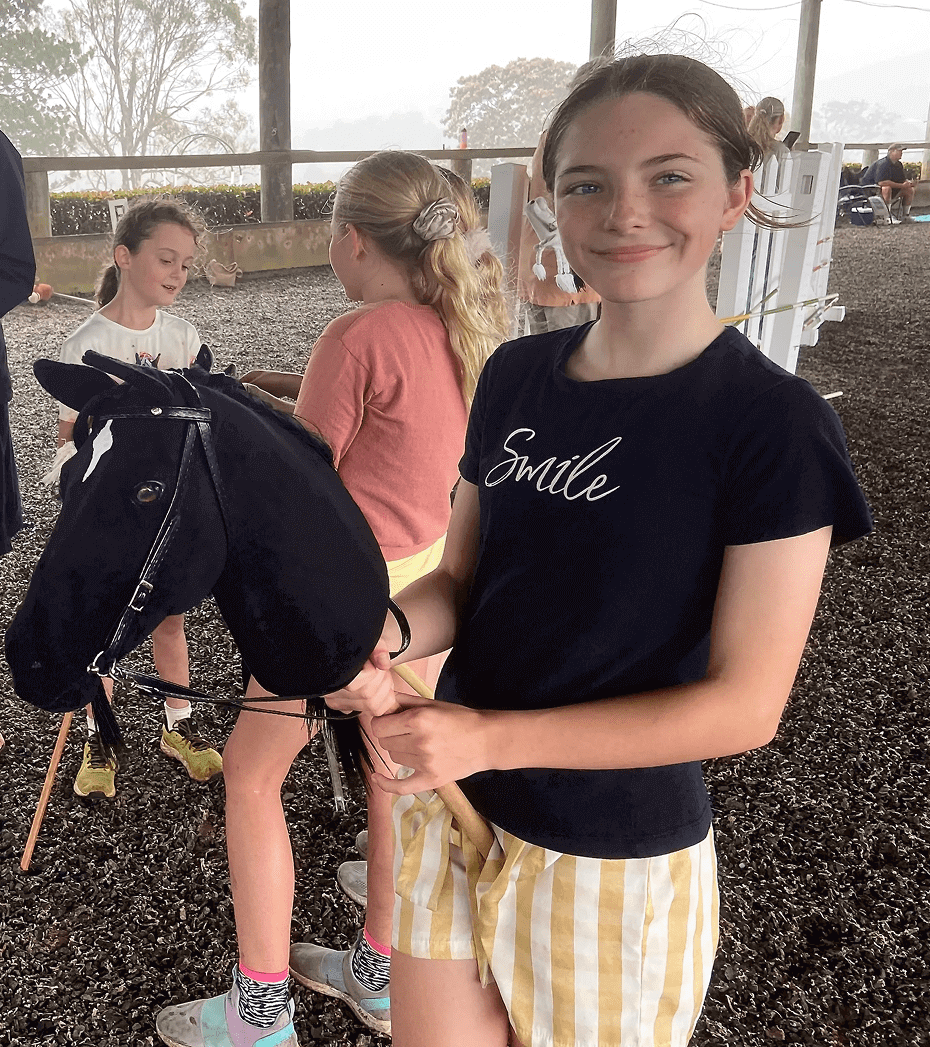 Girl holds a hobby horse, smiling at the camera. She wears a black shirt with "Smile" written on it. Two other girls and equestrian obstacles are in the background.