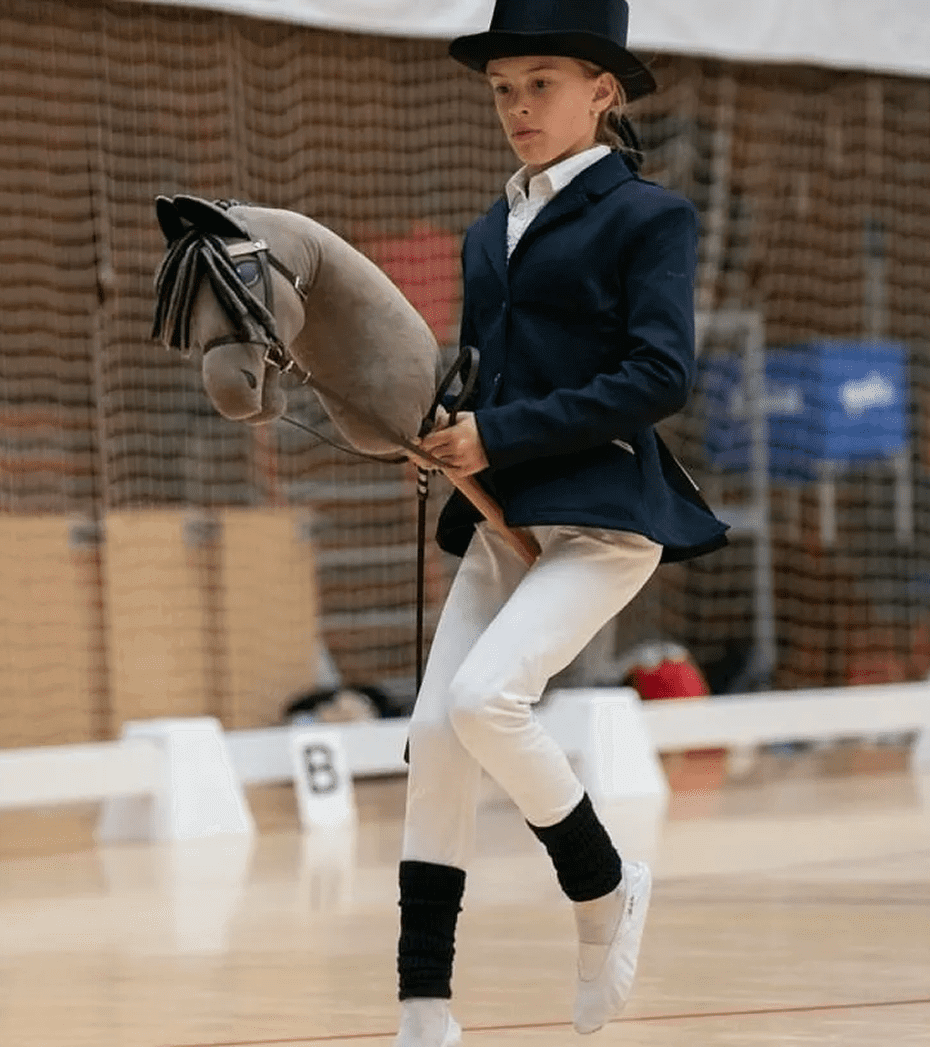 A person in equestrian attire participates in a hobby horse event indoors, holding a toy horse and appearing focused.