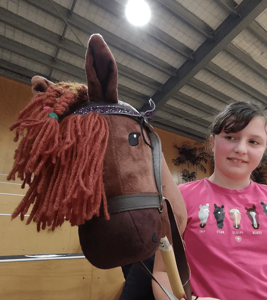 A girl in a pink shirt stands indoors holding a brown hobby horse with a red mane and bridle.