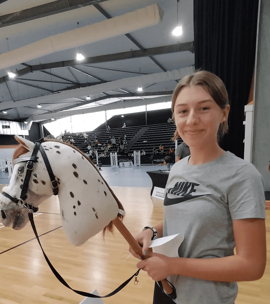 Person holding a hobby horse in an indoor arena with spectators in the background.