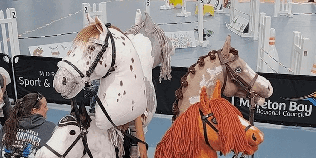 A person holding reins sits on a stick horse next to two others against a backdrop of banners and an indoor arena setup.