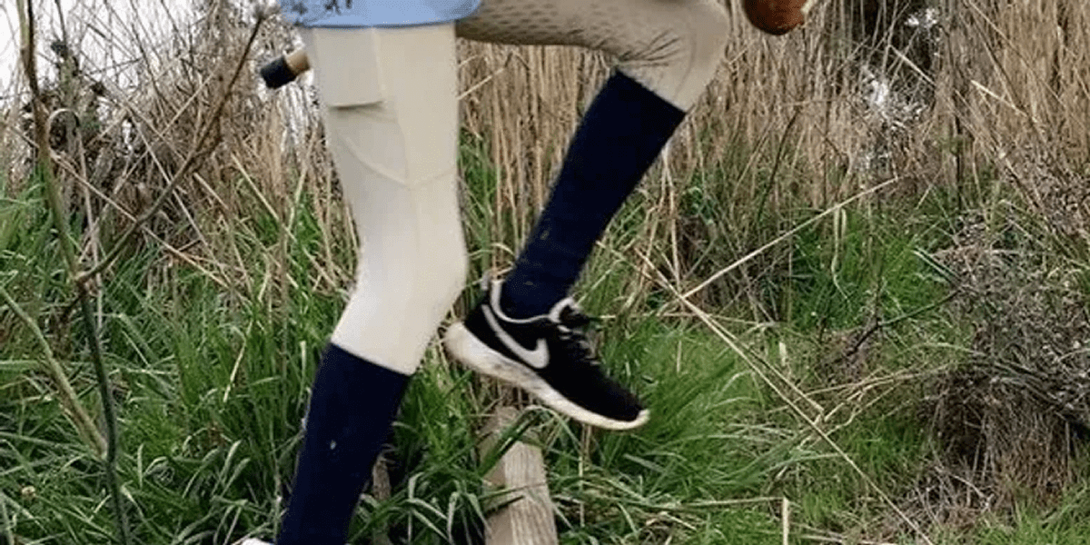 A person in beige pants, blue socks, and black shoes walks on grass. They carry a brown bag and have a knife in a sheath on their thigh.