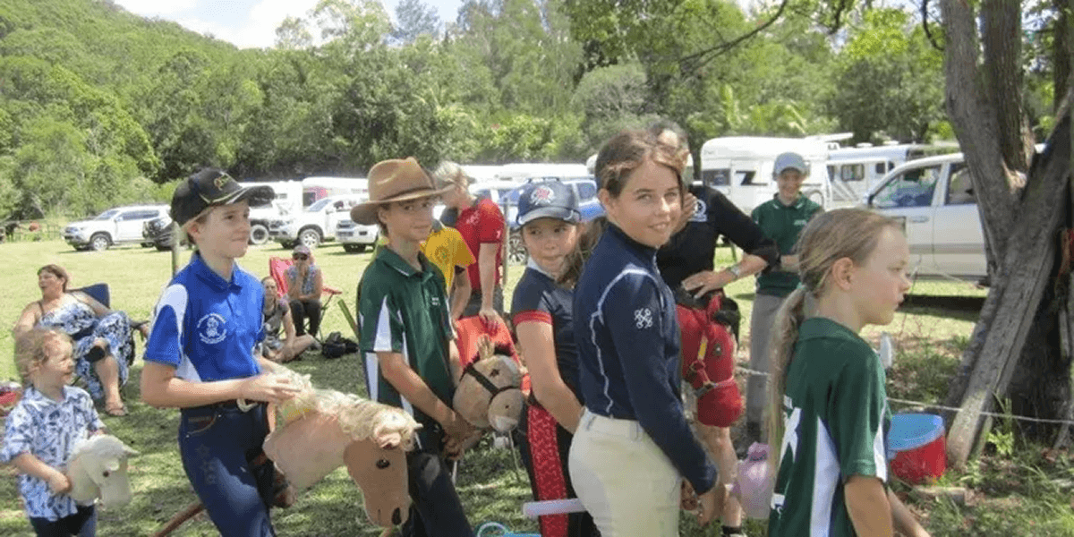 Children participate in a stick horse race outdoors, surrounded by spectators and trees.