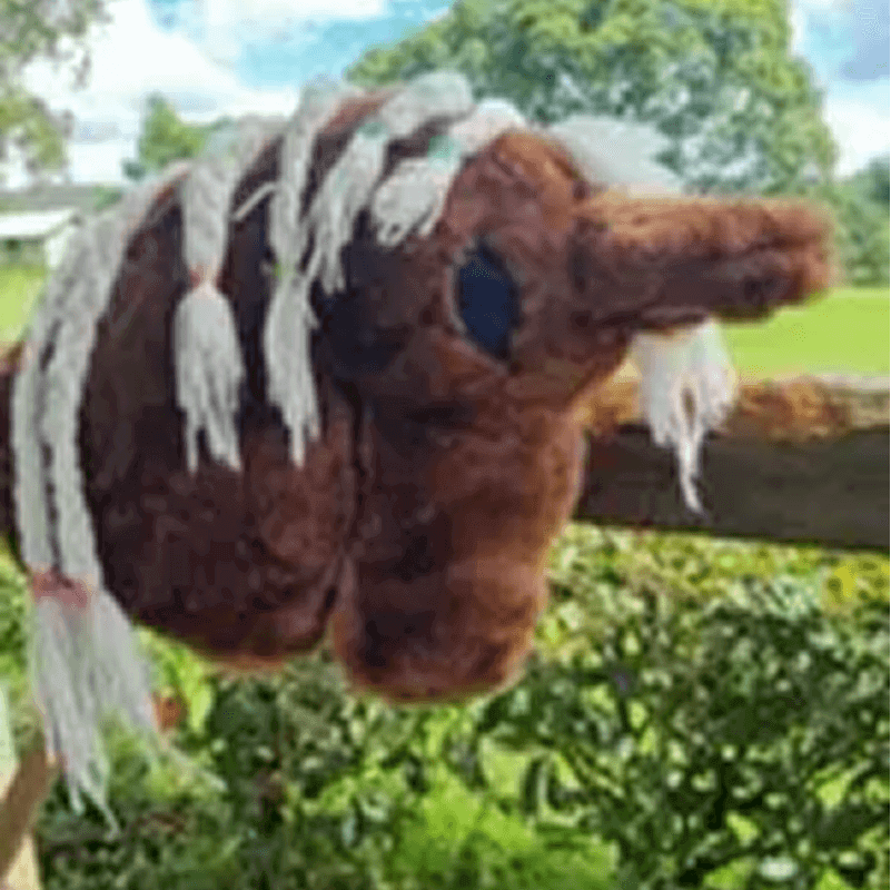 A brown plush hobby horse head with white braided yarn mane, placed on a wooden fence, with greenery and sky in the background.